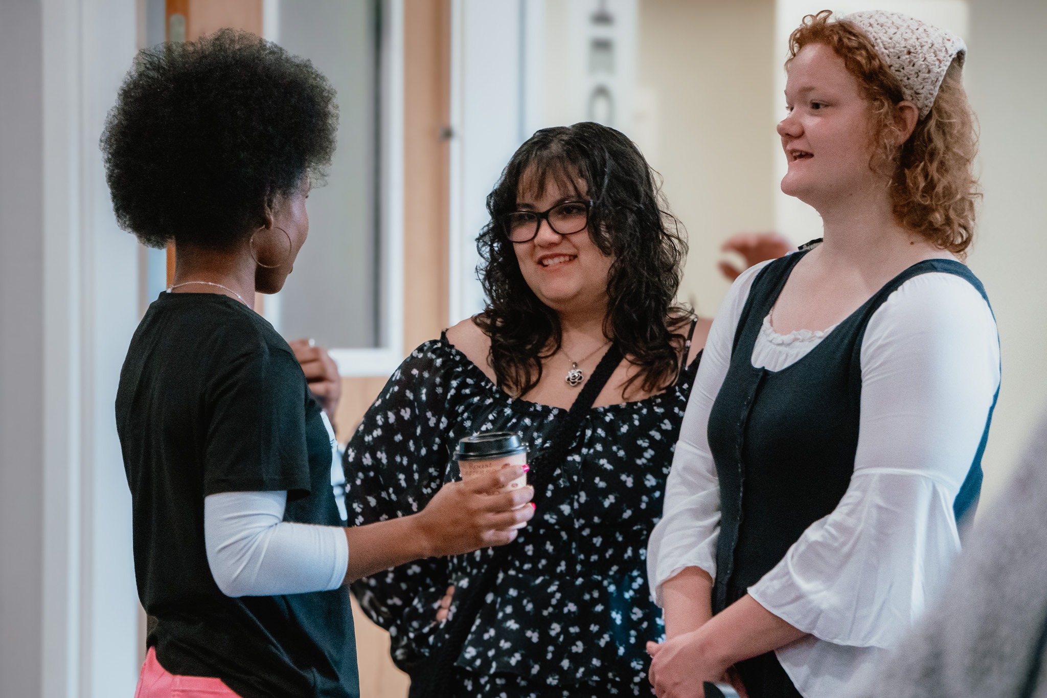 Three Women Engaged In A Friendly Conversation In A Church Lobby. One Woman Holds A Coffee Cup While Speaking, And The Other Two Listen With Smiles. The Setting Is Warm And Welcoming, Reflecting Community And Connection At Church.